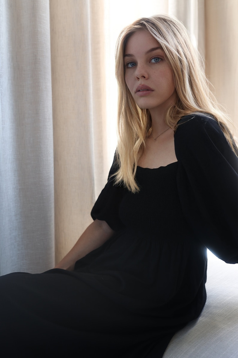 Puede incluir: Una mujer con el pelo largo y rubio lleva un vestido negro de cuello cuadrado con mangas abullonadas. Est&aacute; sentada frente a una ventana con cortinas blancas.