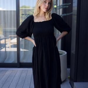 Puede incluir: Una mujer lleva un vestido midi negro con escote cuadrado y mangas abullonadas. El vestido tiene un corpi&ntilde;o fruncido y una falda fluida.