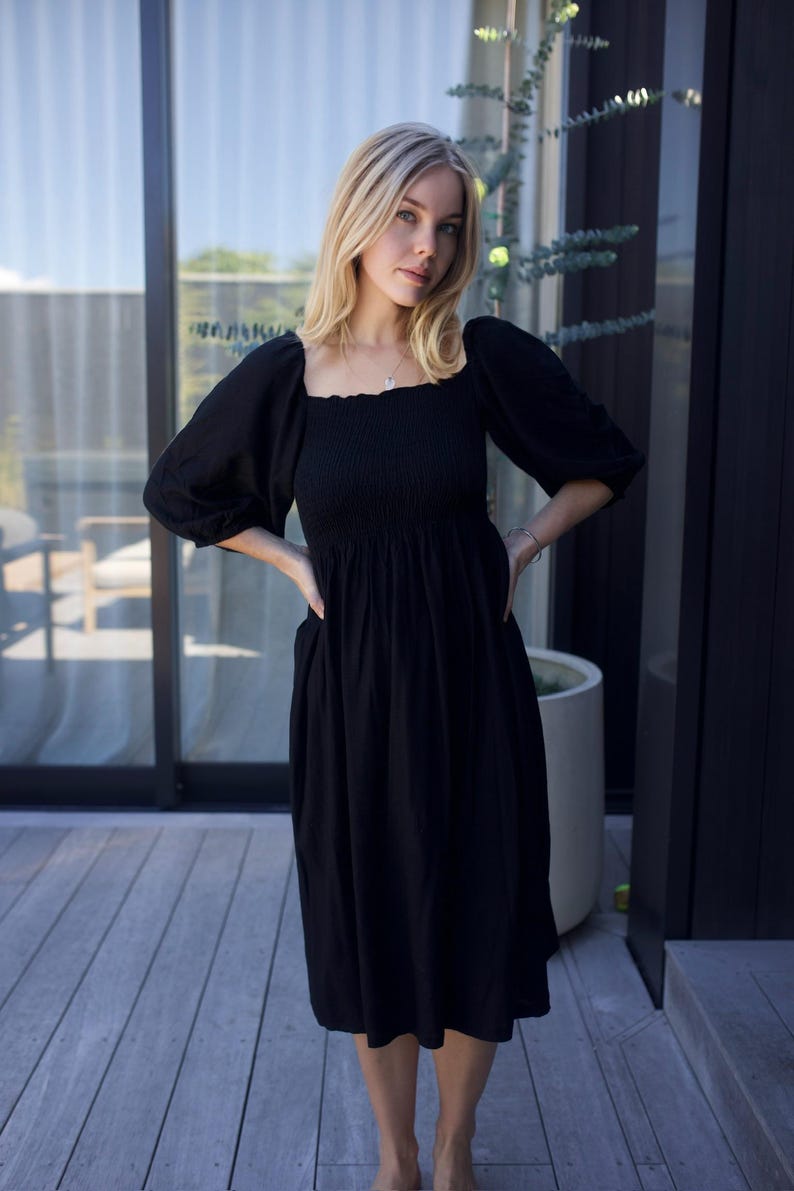 Puede incluir: Una mujer lleva un vestido midi negro con escote cuadrado y mangas abullonadas. El vestido tiene un corpi&ntilde;o fruncido y una falda fluida.