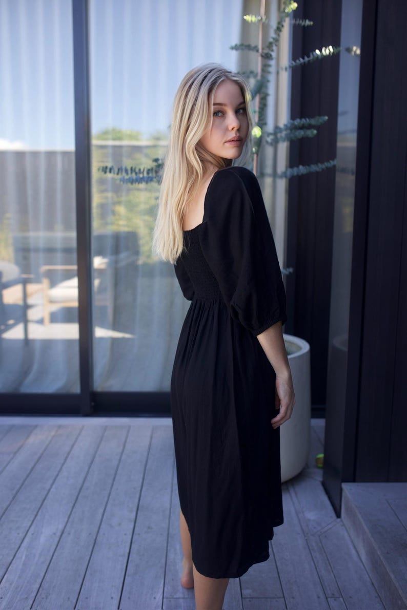Puede incluir: Una mujer con un vestido midi negro de cuello cuadrado y mangas abullonadas. El vestido tiene un corpi&ntilde;o ajustado y una falda fluida.