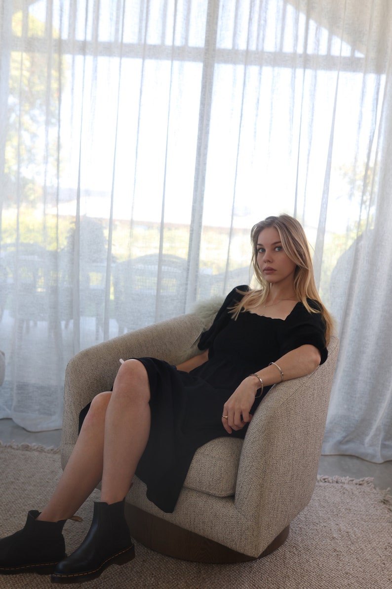 Puede incluir: Una mujer con un vestido negro y botines negros se sienta en un sill&oacute;n gris claro. El sill&oacute;n est&aacute; frente a una ventana con cortinas blancas transparentes.