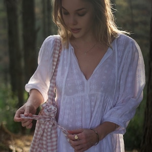 May include: A woman in a white dress with embroidered details is holding a pink and white gingham tote bag. The bag has a drawstring closure and is tied with a pink ribbon.