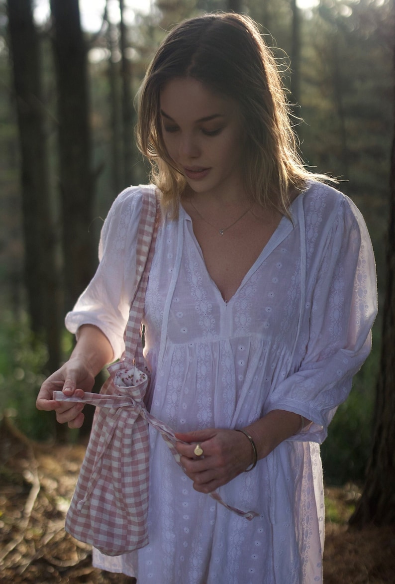 May include: A woman in a white dress with embroidered details is holding a pink and white gingham tote bag. The bag has a drawstring closure and is tied with a pink ribbon.