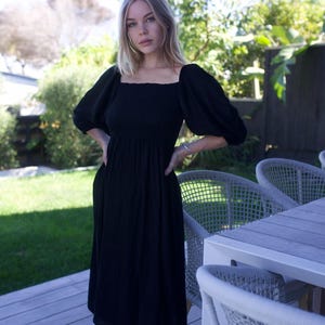 Puede incluir: Una mujer lleva un vestido midi negro con mangas abullonadas y un escote cuadrado. El vestido tiene un corpi&ntilde;o ajustado y una falda fluida. Est&aacute; de pie en una terraza de madera con una silla de mimbre blanca en el fondo.