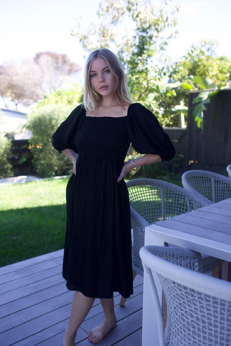 Puede incluir: Una mujer lleva un vestido midi negro con mangas abullonadas y un escote cuadrado. El vestido tiene un corpi&ntilde;o ajustado y una falda fluida. Est&aacute; de pie en una terraza de madera con una silla de mimbre blanca en el fondo.