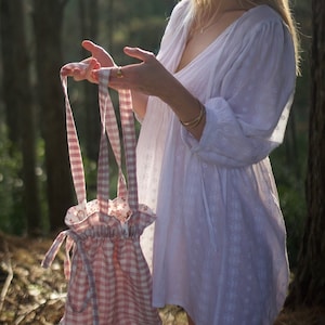 May include: A pink and white gingham drawstring bag with a floral lining. The bag is being held by a person in a white dress.