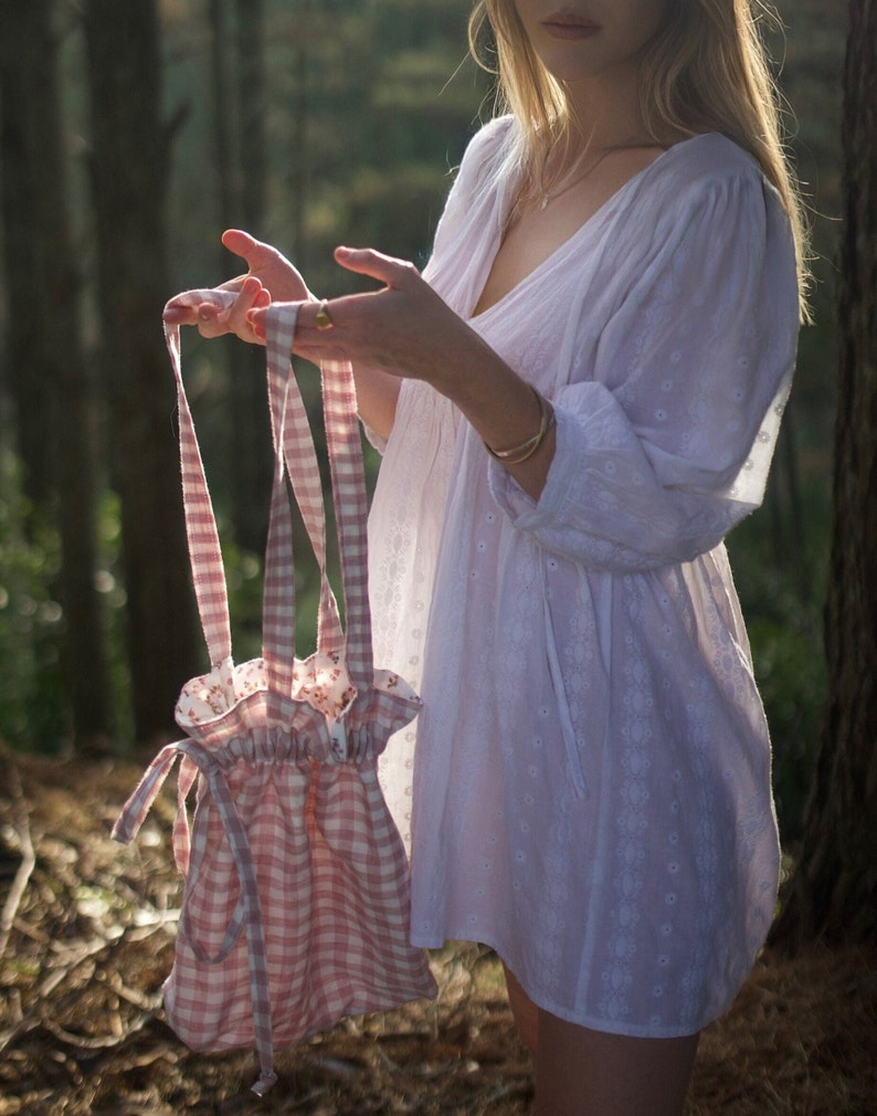 May include: A pink and white gingham drawstring bag with a floral lining. The bag is being held by a person in a white dress.