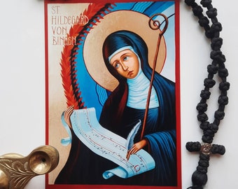 May include: A colorful religious icon depicting Saint Hildegard von Bingen, a German Benedictine abbess, writer, composer, philosopher, Christian mystic, and visionary. The icon features a detailed painting of Saint Hildegard holding a quill and a scroll, with a halo behind her head. The background is a vibrant blue with red and gold accents.