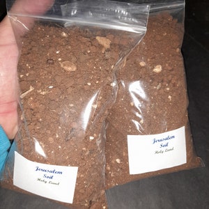 May include: Two clear plastic bags filled with brown soil from Jerusalem, Holy Land. Each bag has a white label with the text "Jerusalem Soil Holy Land".