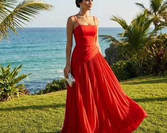 May include: A vibrant red formal gown with a square neckline and spaghetti straps. The dress features a pleated skirt that flows to the ground. A small silver clutch is held. The background includes green grass, palm trees, and a view of the ocean.