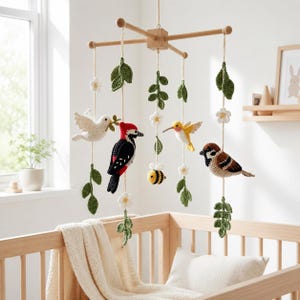 May include: A hanging mobile featuring crocheted birds, including a white dove, a black and red woodpecker, a yellow hummingbird, and a brown sparrow, along with a small yellow and black bee, white flowers, and green leaves suspended from a wooden frame.