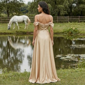 May include: A long, champagne-colored gown with spaghetti straps and off-the-shoulder sleeves. The dress has a flowing skirt and is worn by a person standing near a pond. A white horse grazes in the background near a wooden fence.