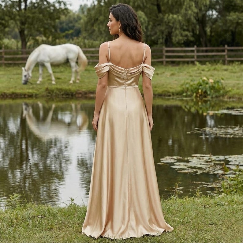 May include: A long, champagne-colored gown with spaghetti straps and off-the-shoulder sleeves. The dress has a flowing skirt and is worn by a person standing near a pond. A white horse grazes in the background near a wooden fence.