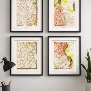 May include: Four framed topographical map prints. Each map is in a black frame with a white mat. The maps feature shades of beige, green, and red, depicting geographical features. The prints are displayed on a light gray wall above a wooden desk.