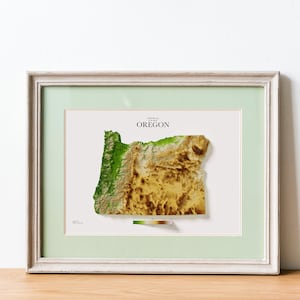 May include: Framed 3D map of Oregon, showcasing the state's topography with green, brown, and tan hues. The map is set against a white background, framed in a light green mat and a white wooden frame. The word "OREGON" is printed at the top.