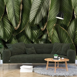 May include: A dark green sofa sits in front of a wall mural featuring large, overlapping tropical leaves with white pinstriped veins. A round wooden coffee table, a potted plant in a white stand, and a black floor lamp complete the arrangement.