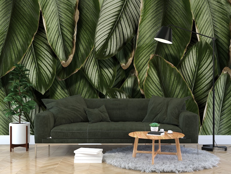 May include: A dark green sofa sits in front of a wall mural featuring large, overlapping tropical leaves with white pinstriped veins. A round wooden coffee table, a potted plant in a white stand, and a black floor lamp complete the arrangement.