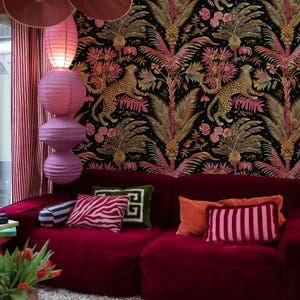 May include: A deep red velvet sofa adorned with various throw pillows in striped, zebra, and solid patterns. The wall behind features black wallpaper with a repeating pattern of leopards, palm trees, and pink flowers, illuminated by stacked paper lanterns.