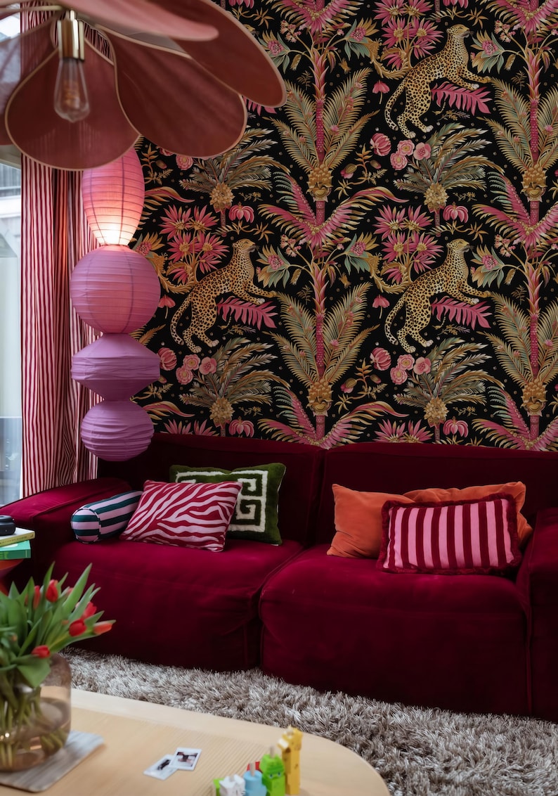 May include: A deep red velvet sofa adorned with various throw pillows in striped, zebra, and solid patterns. The wall behind features black wallpaper with a repeating pattern of leopards, palm trees, and pink flowers, illuminated by stacked paper lanterns.