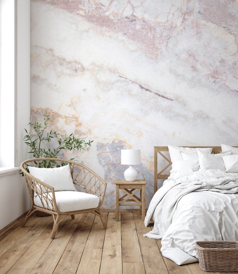 Peut inclure: Une chambre avec un mur au motif de marbre grand format dans des tons de blanc, rose p&acirc;le et gris. La pi&egrave;ce comprend un cadre de lit en bois avec une literie blanche, une petite table de chevet en bois, une lampe blanche et un fauteuil en rotin.