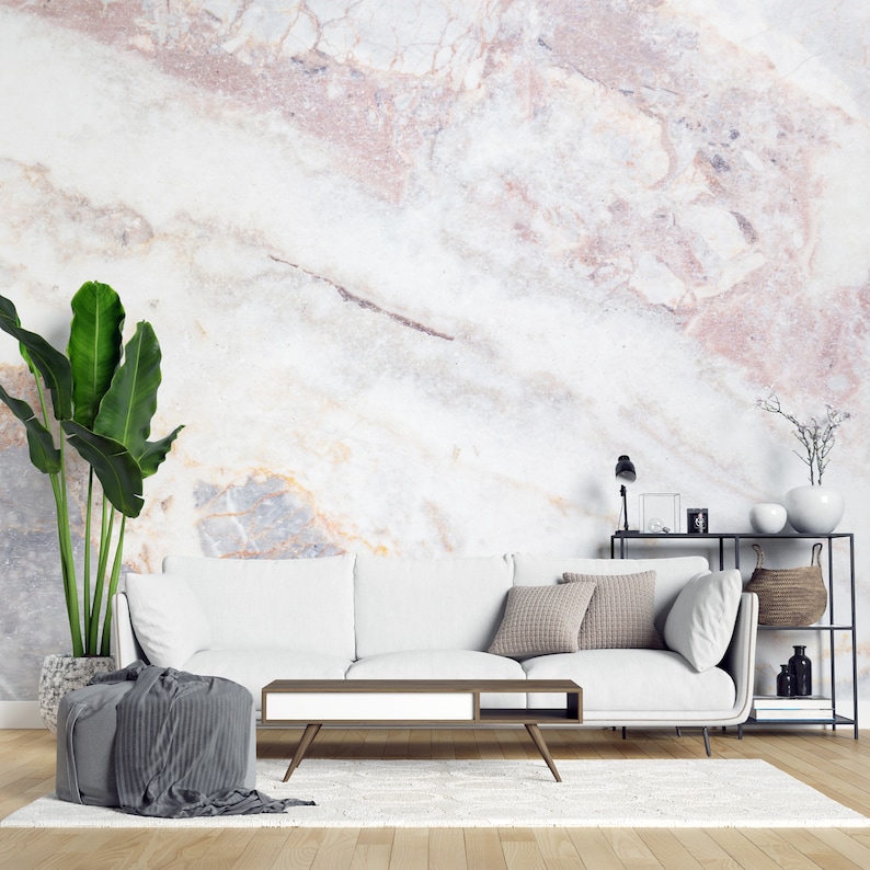 Peut inclure: Un canap&eacute; blanc avec deux coussins gris, une table basse en bois avec un tiroir blanc, un pouf gris avec un tissu drap&eacute;, une grande plante verte dans un pot textur&eacute; et une &eacute;tag&egrave;re en m&eacute;tal noir contre un mur en marbre blanc et rose.