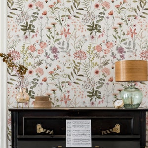 May include: A black upright piano with a sheet of music on the top. The piano is in front of a wall with a floral wallpaper featuring pink and green flowers. A lamp with a wooden base and a glass shade is on the right side of the piano.