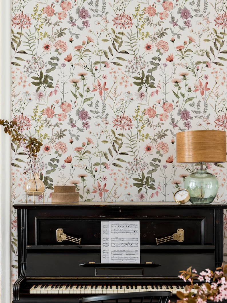 May include: A black upright piano with a sheet of music on the top. The piano is in front of a wall with a floral wallpaper featuring pink and green flowers. A lamp with a wooden base and a glass shade is on the right side of the piano.