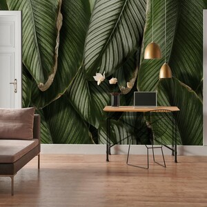 May include: A wall mural featuring large, overlapping dark green tropical leaves with visible veins. The mural covers the wall behind a wooden desk with a laptop, a vase with white flowers, and two gold pendant lights, flanked by two white doors.