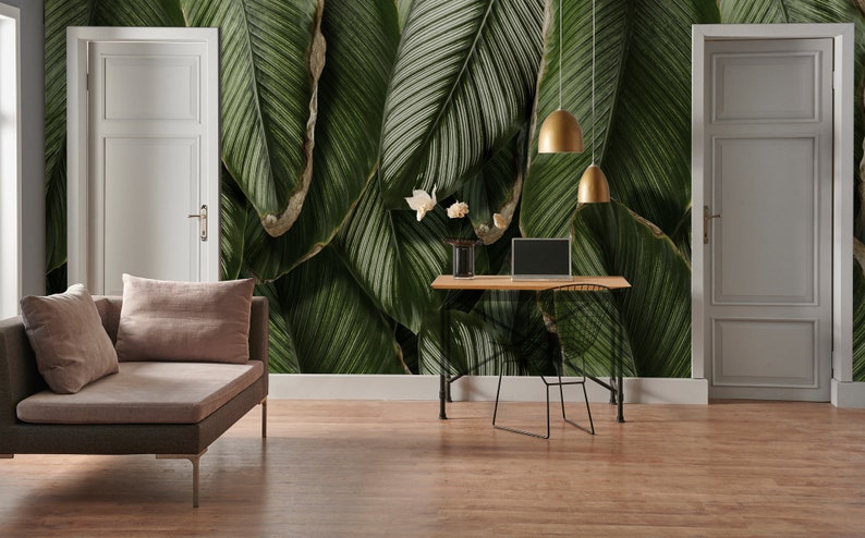May include: A wall mural featuring large, overlapping dark green tropical leaves with visible veins. The mural covers the wall behind a wooden desk with a laptop, a vase with white flowers, and two gold pendant lights, flanked by two white doors.