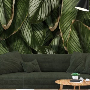 May include: A dark green sofa sits before a wall mural featuring large, overlapping tropical leaves with white pinstriped veins. A wooden coffee table holds a small succulent, a black book, and two white cups, with a gray rug underneath.