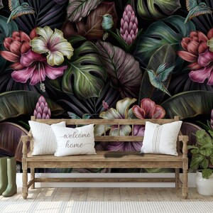 May include: A wooden bench with two white pillows, one featuring the text "welcome home", placed against a dark wallpaper mural of tropical leaves, pink hibiscus flowers, and hummingbirds. Olive green boots and a potted plant sit nearby.