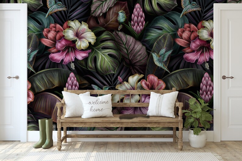 May include: A wooden bench with two white pillows, one featuring the text "welcome home", placed against a dark wallpaper mural of tropical leaves, pink hibiscus flowers, and hummingbirds. Olive green boots and a potted plant sit nearby.