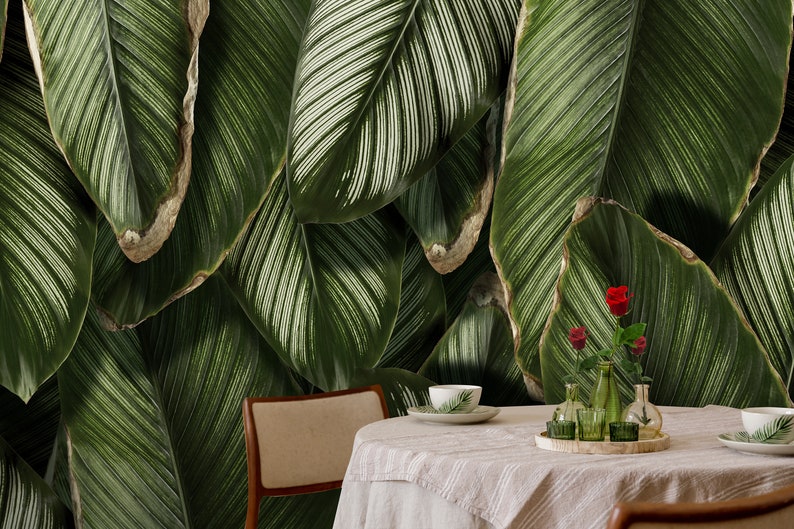 May include: A dining table with a light-colored tablecloth, two chairs, and a wooden tray holding three small glass vases with red roses and green leaves. The background features a large-scale wall mural of dark green tropical leaves.