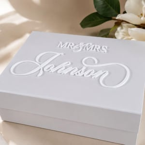 May include: A rectangular white gift box with a lid featuring raised white lettering. The text on the lid reads MR & MRS in a simple font, followed by the name Johnson in a large, elegant, cursive script. The box has a smooth, matte finish.