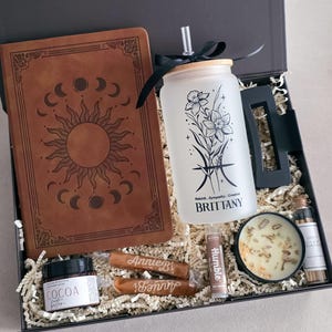 May include: A gift box containing a brown leather-look journal with a sun and moon design, a frosted glass tumbler with a bamboo lid, straw, and floral graphic with the name BRITTANY, a black hair clip, cocoa butter, two Annie B's treats, lip balm, a candle, and matches.