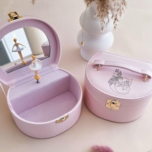 May include: Two light pink jewelry boxes with gold-tone latches. One box is open, revealing a lavender interior, a round mirror, and a rotating ballerina figurine. The other box is closed, featuring a top handle and a floral letter C engraving.