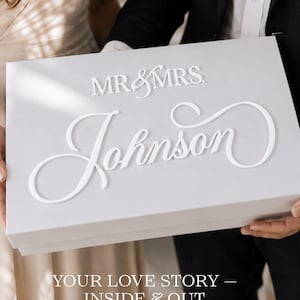 May include: A rectangular light gray gift box featuring the white raised text MR & MRS and the name Johnson in a cursive script. The box has a clean, smooth finish and is held horizontally, displaying the personalized lettering on the front lid.