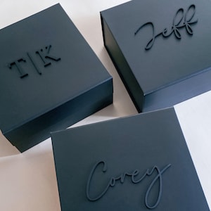 May include: Three dark blue gift boxes with custom names and initials. One box has the initials "T|K", another has the name "Jeff", and the third has the name "Corey" in a cursive font. The boxes are arranged on a light-colored surface.