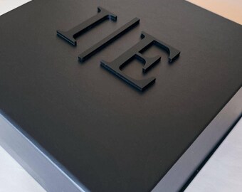 May include: A square black box with a matte finish featuring the raised, three-dimensional black letters I and E centered on the lid. The box has a clean, structured design with sharp edges and a solid, uniform dark color throughout the exterior.