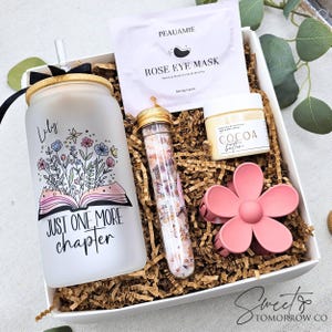 May include: A gift box with a frosted glass tumbler with a floral design and the text "Just One More Chapter." The box also contains a rose eye mask, cocoa butter, a pink flower hair clip, and a tube of bath salts.