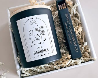 Puede incluir: Una vela negra con tapa de madera y una etiqueta con un diseño de constelación de Leo y el nombre "Barbara". La vela está en una caja blanca con papel decorativo y una caja de cerillas que dice "Sweet Tomorrow Co premium matches."