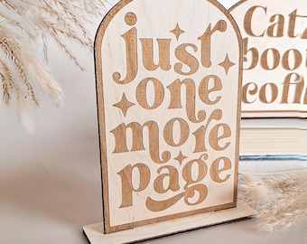 Könnte beinhalten: Ein gewölbtes Holzschild mit heller, natürlicher Oberfläche und dem lasergravierten Text "just one more page" in einer stilisierten Schriftart, akzentuiert durch kleine vierzackige Sterne. Das Schild steht auf einem rechteckigen Holzsockel.