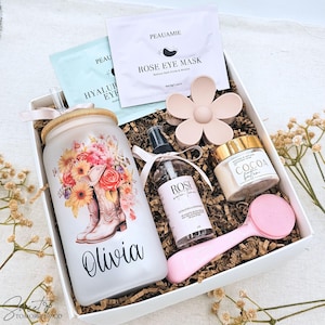 May include: A white gift box containing a frosted glass tumbler with a floral cowboy boot design and the name Olivia, a rose water toner spray, a cocoa butter jar, a pink silicone facial brush, a flower hair clip, and two eye mask packets.