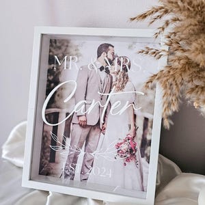 May include: A white shadow box frame containing a photograph of a couple kissing. The glass features white text reading MR. & MRS. Carter, with a decorative leaf graphic and the text EST. 2024 printed below the couple in a script and serif font.