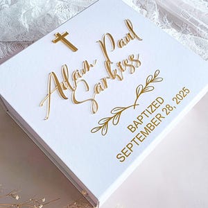 May include: A white square keepsake box featuring gold lettering that reads Adam Paul Sarkiss, a gold cross icon, and gold text stating BAPTIZED SEPTEMBER 28, 2025. The lid is decorated with a gold line art branch design and a small gold cross.
