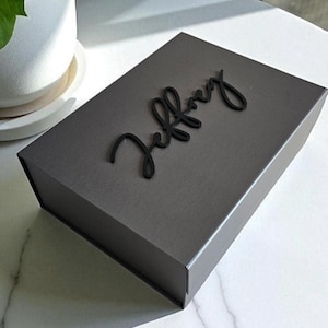 May include: A rectangular dark gray gift box with a matte finish. The top of the box features the name Jeffrey in a raised, black, cursive script font. The box has a clean, structured design with sharp edges and a solid, uniform color throughout.