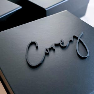 May include: A square black gift box featuring the name Corey in raised black script lettering attached to the lid. The box has a smooth matte finish and a hinged lid design. Another similar black box with a name is partially visible in the back.