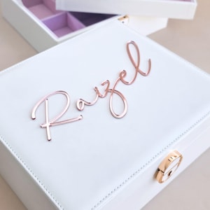 May include: A white square jewelry box with a rose gold metallic latch and the name Razel in rose gold script lettering on the lid. The box features stitched edges and is shown with an open matching white jewelry tray with light purple lining.