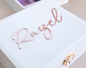 May include: A white square jewelry box with a rose gold metallic latch and the name Razel in rose gold script lettering on the lid. The box features stitched edges and is shown with an open matching white jewelry tray with light purple lining.