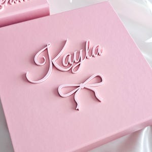 May include: A square light pink gift box featuring the name Kayla in raised, script-style lettering and a small, matching pink bow icon attached to the lid. A second, partially visible pink box with the name Kendall is positioned above it.